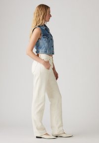 Γυναίκα με αμάνικο μπλε τζιν γιλέκο, ψηλόμεσα ολόλευκα ίσια τζιν παντελόνια και λευκά φλατ παπούτσια, στέκεται προφίλ μπροστά σε απλό φόντο.