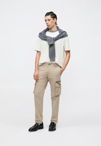 Καθημερινό ντύσιμο με ανοιχτό μπεζ μπλουζάκι, μπεζ παντελόνι cargo με πλαϊνές τσέπες και γκρι πλεκτή ζακέτα που φοριέται γύρω από το λαιμό. Μαύρα παπούτσια.