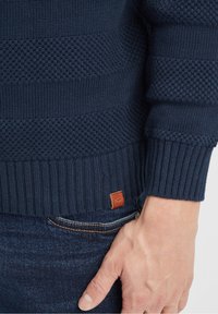 Maglione lavorato a maglia blu navy con strisce testurizzate, con polsini a coste e un'etichetta in pelle marrone sul bordo inferiore, abbinato a jeans denim scuri.