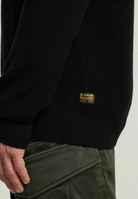 Maglione nero lavorato a maglia con polsini e orlo a coste, dotato di una patch con il logo G-Star RAW sul lato. La texture sembra calda e resistente.