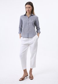 Camicia grigia con bottoni, leggermente strutturata, maniche lunghe e un piccolo logo. Abbinata a pantaloni bianchi fluidi e sandali con tacco marroni.