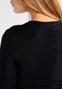 Svart figursydd topp med slät textur, rund halsringning och en diskret etikett nära axeln. Minimalistisk design med långa ärmar.