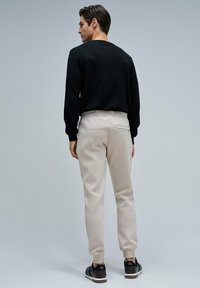 Hombre de pie de espaldas, vistiendo una camiseta negra de manga larga, pantalones jogger beige y zapatillas negras frente a un fondo gris liso.