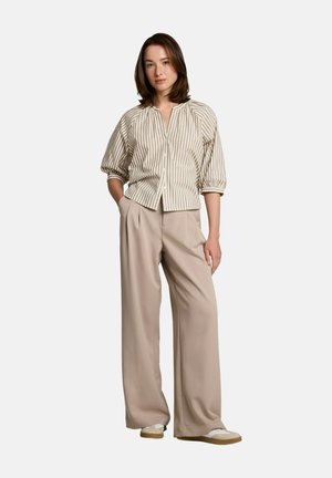 Vrouw staat, draagt beige wijde broek, beige-wit gestreept blouse met knopen en pofmouwen, en lichtgekleurde sneakers.