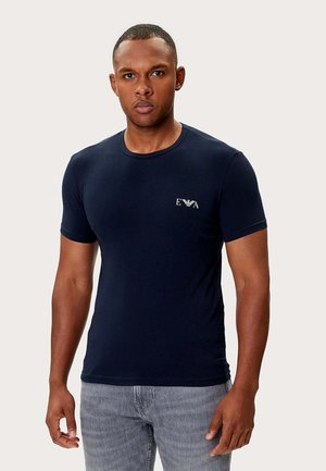 Uomo che indossa una maglietta aderente blu navy con logo bianco "EVA" sul petto e jeans grigio chiaro, in piedi contro uno sfondo chiaro e uniforme.