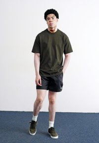 Olivenfarvet t-shirt med korte ærmer og subtilt logo, parret med sorte mønstrede shorts og lysegrå sokker, afsluttet med mørkegrønne sneakers.