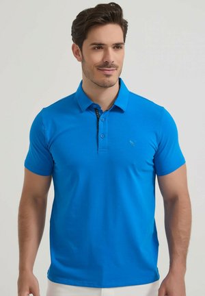 Homme portant un polo bleu vif à manches courtes, debout devant un fond clair uni, regardant légèrement sur le côté.
