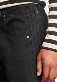 Černé džíny z denimu s úzkým střihem, jemnou texturou a stříbrnými detaily. Je na sobě stylový dlouhý rukáv s pruhy, který je částečně vidět.