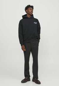 Sudadera negra con un logo en la parte frontal, combinada con pantalones oscuros a medida y zapatos marrones de cordones. Textura de tela suave y corte relajado.