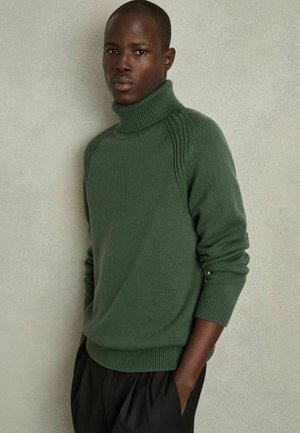 Pulover verde cu guler înalt, cu o textură canelată și detalii pe umeri. Purtați cu pantaloni negri, largi. Design simplu, modern.