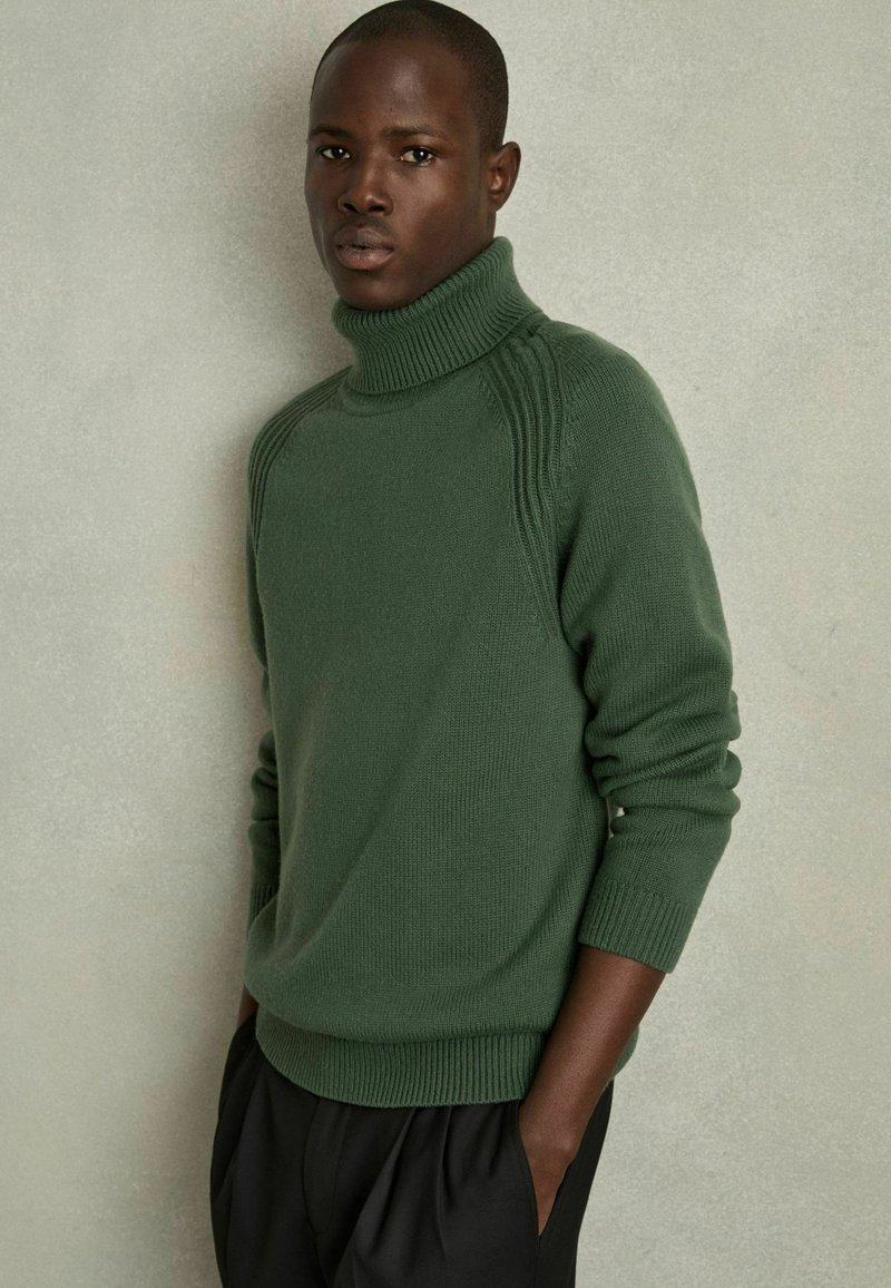 Pulover verde cu guler înalt, cu o textură canelată și detalii pe umeri. Purtați cu pantaloni negri, largi. Design simplu, modern.