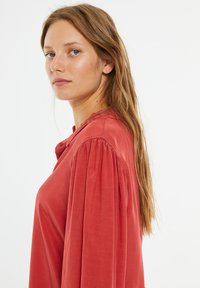 Blusa roja con un corte relajado, que presenta mangas abullonadas y un detalle de botones en el cuello. Tejido suave con una textura lisa.