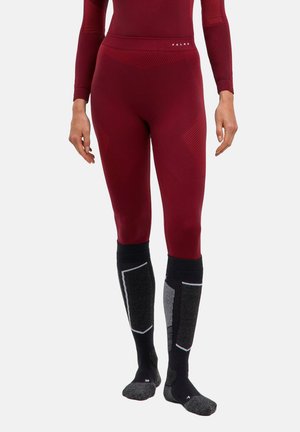 Kvinde iført sømløse burgunderrøde leggings og sorte knæhøje polstrede sokker, stående foran en ensfarvet baggrund.