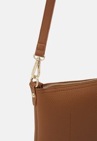 Hnědá kožená taška s texturovaným povrchem, která disponuje zipem a kovovými sponami ve zlatém tónu. Navržena v elegantním obdélníkovém tvaru.