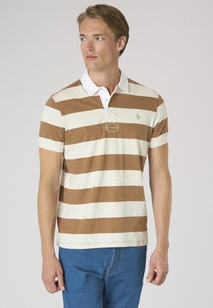 Giovane uomo che indossa una polo a righe marroni e bianche e jeans blu, guarda a sinistra davanti a uno sfondo uniforme.