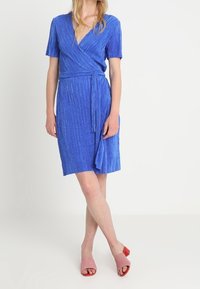Modrá zavinovací šaty s krátkými rukávy, vertikální texturou a uvázaným pasem. Nosené s růžovými sandály na podpatku s otevřenou špičkou.