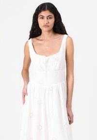 Vestito bianco senza maniche con scollatura quadrata, vita arricciata e motivi floreali ricamati su tutta la gonna. Materiale in cotone.