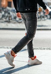 Tummat farkut, joissa maalisirotuksia, käännetyt lahkeet, ja harmaat lenkkarit punaisilla yksityiskohdilla kävelemässä vaalealla jalkakäytävällä.