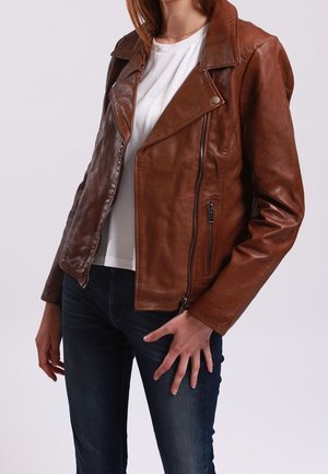 Femme portant une veste en cuir marron sur un t-shirt blanc et un jean bleu foncé, debout avec une main près de sa hanche.