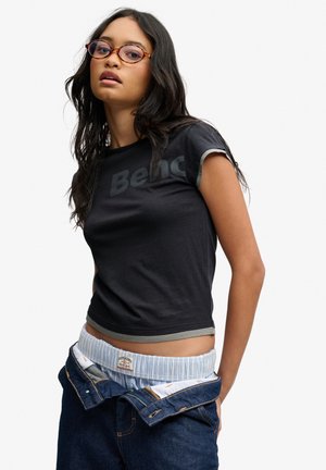 Junge Frau mit langen dunklen Haaren, die Schildpattbrille, ein schwarzes kurzes Oberteil und aufgeknöpfte Jeans trägt, die den gestreiften Bund der Unterwäsche offenbaren.