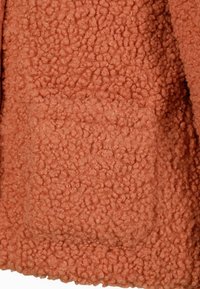 Textúrált, bolyhos anyag meleg terracotta színben, egyetlen zsebbel és puha, gyapjas megjelenéssel.