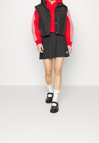 Czarna kamizelka puchowa na czerwonym hoodie z białymi paskami, w połączeniu z plisowaną czarną spódnicą, białymi skarpetkami i czarnymi butami na klamrę.