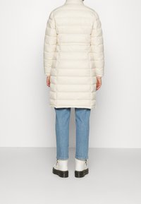 Kremowa puchowa kurtka z wysokim kołnierzem, z pikowanymi sekcjami, bocznymi rozcięciami i długością do kolan, w połączeniu z jasnoniebieskimi dżinsami i białymi botkami.