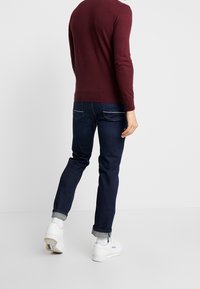 Burgundy stickad tröja, denimjeans och vita sneakers. Tröjan har ribbade mudd och fåll; jeansen är mörka med synlig sömnad.
