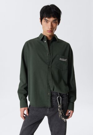 Young man wearing dark green button-up shirt with "ATHLETIX DEPT" on pocket, black jeans, and keychain hanging from belt loop.