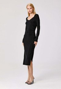 Femme portant une robe midi noire à manches longues ajustée avec une fente sur le côté, associée à des sandales à talons hauts noires avec des brides transparentes.