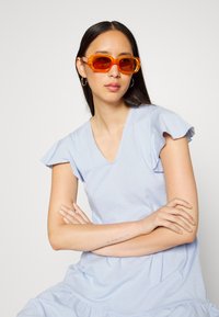 Vestido de algodão azul claro com mangas curtas e franzidas e um decote em V. A modelo usa óculos de sol retangulares laranja e brincos de argola prateados simples.