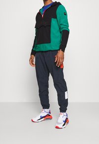 Felpa con cappuccio verde e nera con tasca a marsupio; abbinata a pantaloni blu navy con blocchi di colore e accenti bianchi, completati da sneakers bianche.