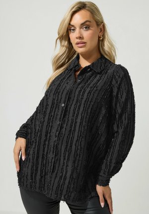 Blonde vrouw die een gestructureerd zwart overhemd met lange mouwen, knopen en verticale strepen draagt en een zwarte broek tegen een effen achtergrond.