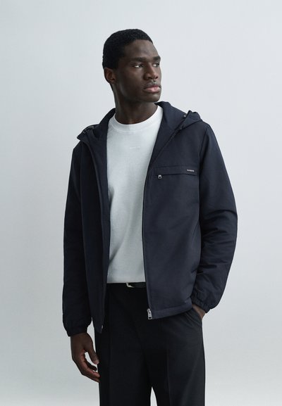 Jeune homme portant une veste à capuche noire sur une chemise blanche, debout avec une main dans la poche, regardant sur le côté sur un fond uni.