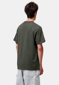 Hombre con cabello corto y oscuro, vestido con una camiseta verde oscuro de gran tamaño y pantalones cortos cargo gris claro, de pie de espaldas sobre un fondo blanco.