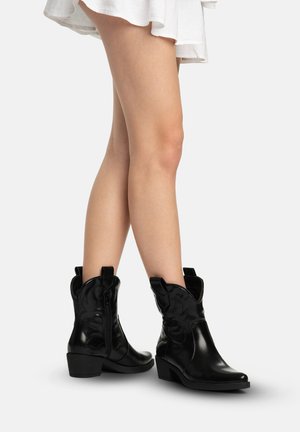 Ben iført kort hvid nederdel og sorte læder cowboystøvler til midt på læggen med dekorative syninger og lave hæle på neutral baggrund.