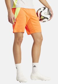 Heldere oranje sportshorts met een gaaspaneel, gecombineerd met witte voetbalschoenen en knielange witte sokken, met in de hand een geconfigureerde voetbal.