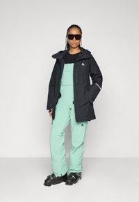 Chaqueta impermeable negra con capucha, combinada con un mono verde menta. La modelo lleva gafas de sol y botas de esquí, mostrando un diseño orientado al deporte.
