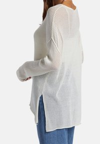 Leichter, gestrickter Pullover aus Creme mit vertikalen Rippstrukturen, langen Ärmeln und seitlichen Schlitzen. Lockere Passform, leicht durchscheinende Textur.