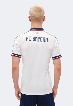 Osoba blondynka ubrana w biały trykot FC Bayern z granatowymi i czerwonymi wykończeniami, stojąca tyłem do widza.