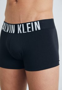 Sorte bomullsbokser med en bred elastisk midjebånd som har "CALVIN KLEIN" i fet hvit skrift. Glatt tekstur og tettsittende design.