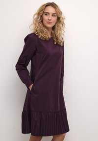 Violetti puuvillamekko pitkillä hihoilla, pleidatareuna ja kaksi sivutaskua. Sileä rakenne ja rento istuvuus, yltää polven ylle.
