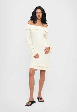Off-the-shoulder ribbed dress in ivory with ruffled edges, long sleeves, and a fitted shape. Paired with black flip-flops.