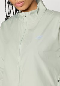 Ανοιχτό πράσινο μπουφάν με φερμουάρ, κατασκευασμένο από ελαφρύ ύφασμα, διαθέτοντας ψηλό γιακά και ανάγλυφο λογότυπο στο στήθος. Λείος υφή.