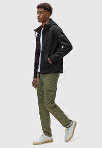 Giacca con zip nera e cappuccio, in tessuto leggero, con tasche frontali; abbinata a pantaloni cargo color oliva e sneaker bianche.