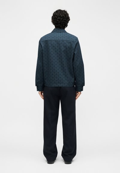 Άνδρας με σγουρά μαλλιά που φοράει σκούρο σακάκι με σχέδια και φαρδιά μαύρη παντελόνα, στέκεται γυρισμένος πλάτη σε απλό λευκό φόντο.