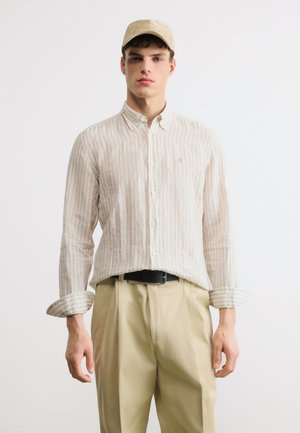 Νεαρός άνδρας που φοράει μπεζ ριγέ πουκάμισο με κουμπιά, μπεζ παντελόνι, μαύρη ζώνη και μπεζ καπέλο, στέκεται μπροστά σε λευκό φόντο.