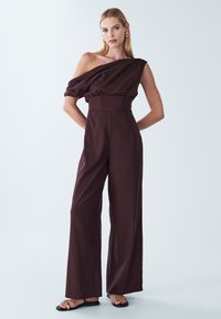 Brązowy kombinezon z jednym ramiączkiem, z bufiastym rękawem, paskiem w talii i szerokimi nogawkami. Gładka tekstura, minimalistyczny styl i elegancka sylwetka.