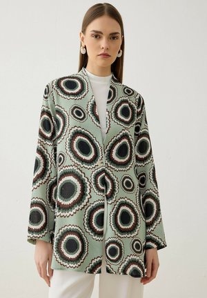 Femme aux cheveux bruns raides porte un blazer vert à grands motifs floraux circulaires sur un haut blanc et un pantalon, debout contre un mur uni.