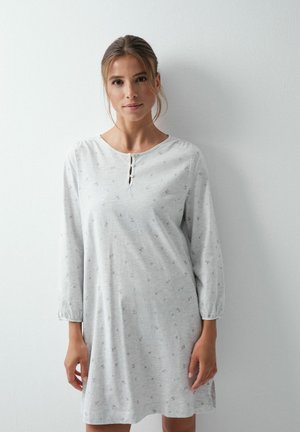 Une femme présente, devant un mur uni, une chemise de nuit gris clair à manches longues, ornée d’un petit motif floral et dotée d’une encolure boutonnée.